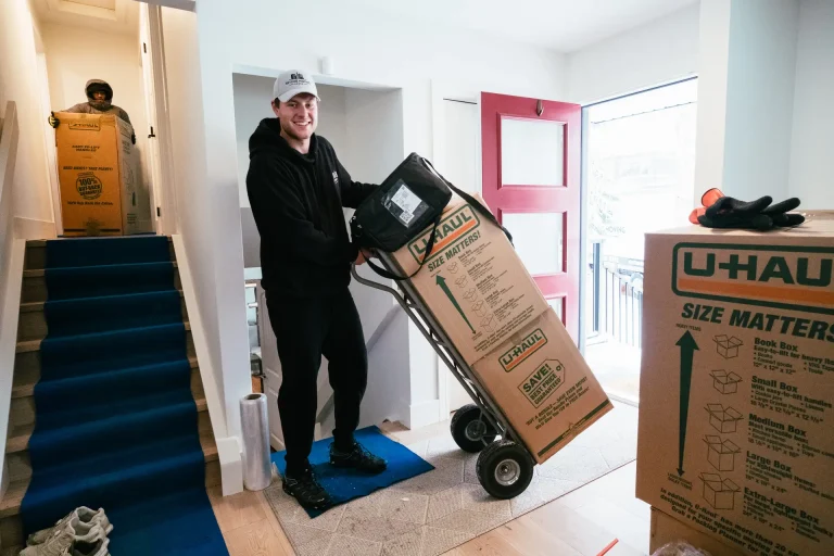 A person is wheeling a large moving box through a doorway, while another person carries a box nearby